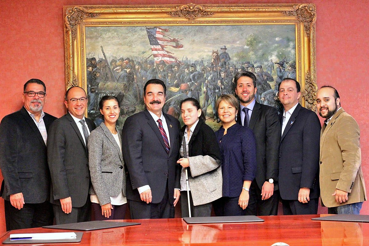 Priscilla Garces, holding a white cane, stands at the center of a group of ten civic and community leaders in front of a large historical painting.