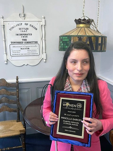 Priscilla Garces, con chaqueta rosa, sostiene un reconocimiento enmarcado del Municipio de Union, Celebrating Women, de marzo de 2023, que la nombra Disability Advocate and Climate Activist y SHEro Advocate. Su bastón blanco descansa a su lado.