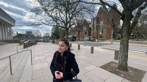 Priscilla Garces, con abrigo azul marino, sonríe de pie en una plaza de piedra, con árboles sin hojas y edificios de ladrillo detrás.