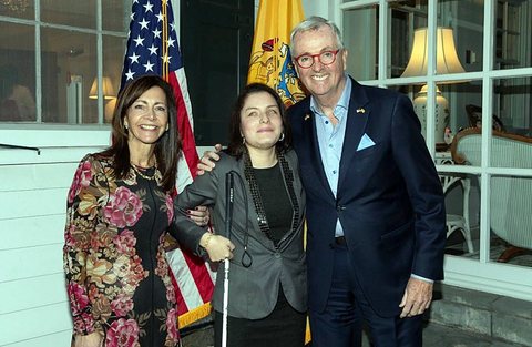 Priscilla Garces, con su bastón blanco, de pie entre el gobernador de Nueva Jersey, Philip D. Murphy, y otro invitado, delante de las banderas de Estados Unidos y de Nueva Jersey.