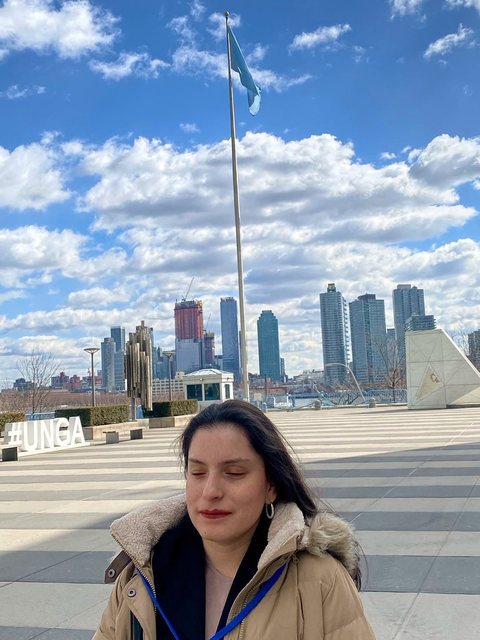 Priscilla Garces, con abrigo de invierno y una acreditación azul, de pie en la plaza de la Sede de las Naciones Unidas, con la bandera de la ONU, el cartel #UNGA y el perfil de Manhattan detrás.